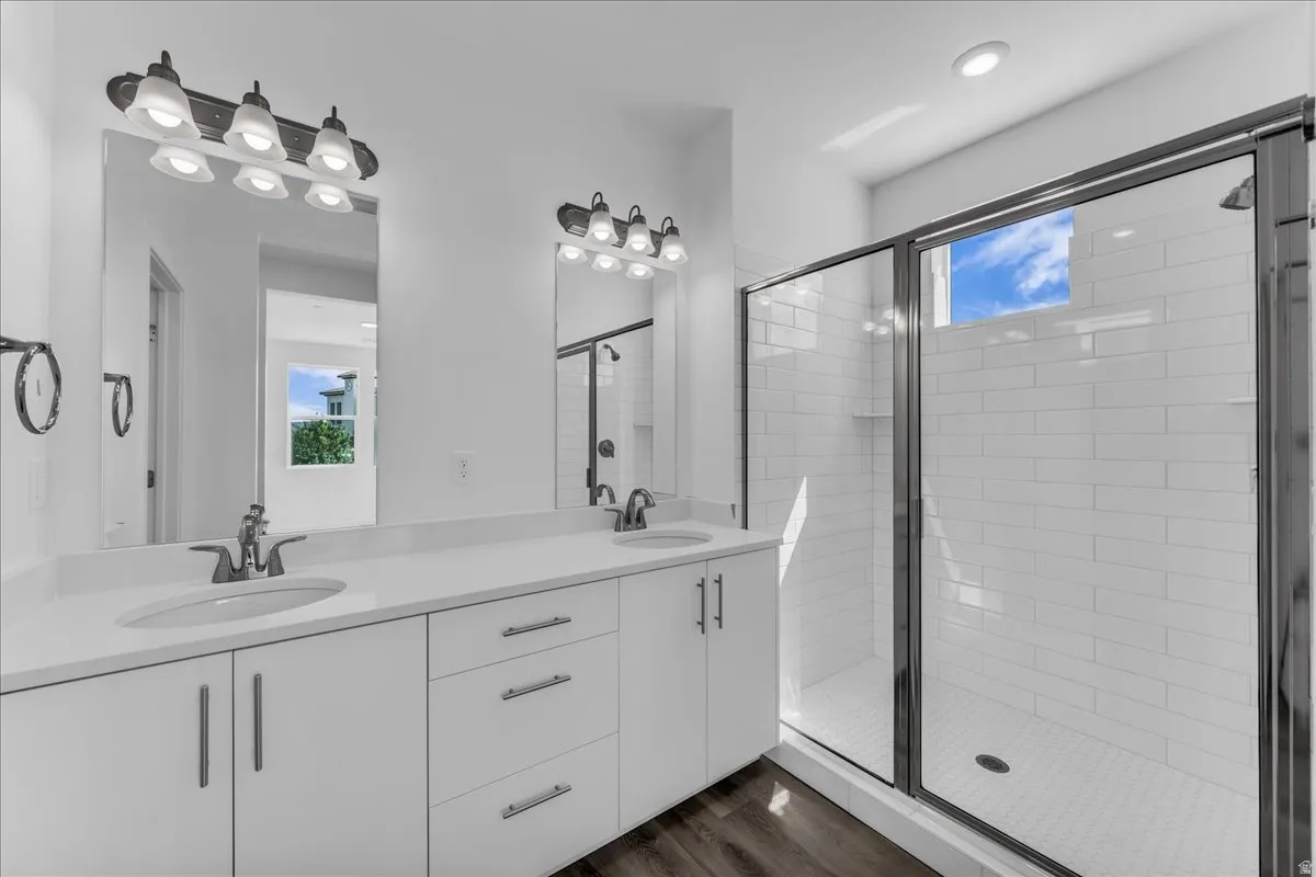 Double vanity with white cabinetry and integrated sinks, a walk-in shower with clear glass enclosure, subway tile shower surround, and wood-finish flooring