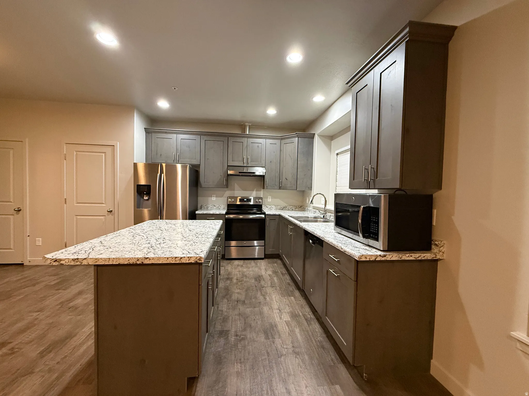 Kitchen featuring wood-finish flooring, shaker-style cabinetry, granite countertops, a stainless steel appliance suite, and an island with bar seating