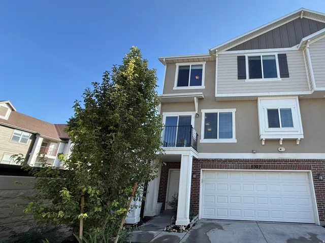 Three-story exterior featuring a mix of siding and stucco, brick detailing, a private balcony, an attached two-car garage, and a manicured front yard with mature landscaping
