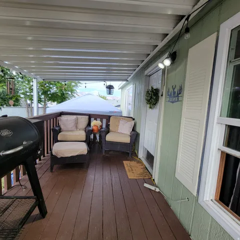 Covered wood-finish deck with a wood railing, leading to a light green exterior with a white entry door and white window shutters