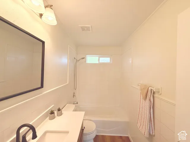 White tiled shower with a showerhead and tub, wood-finish flooring, white vanity with a rectangular sink and dark-toned faucet, wall-mounted mirror, and overhead lighting fixture