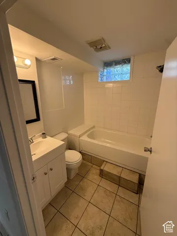 White vanity with an integrated sink, tiled flooring, bathtub with white tile surround, and a glass block window