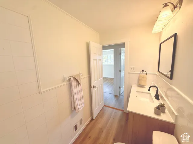 Wood-finish flooring, white tile wainscoting, white vanity with integrated sink, black wall-mounted faucet, and rectangular framed mirror