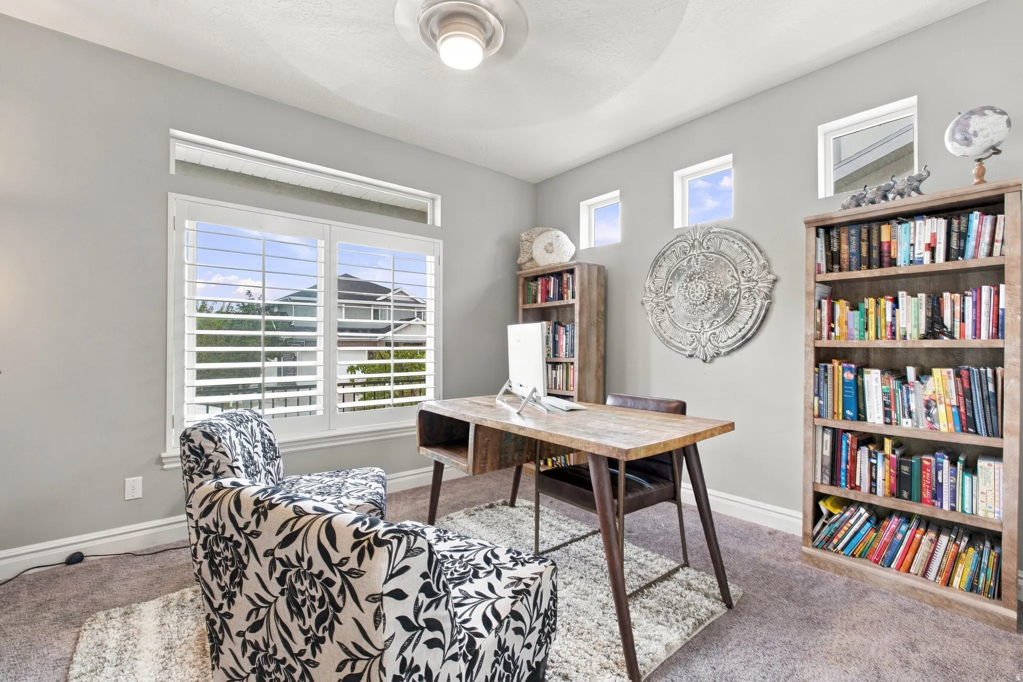 Neutral wall paint, ceiling fan with light fixture, multiple windows with white plantation shutters, built-in shelving