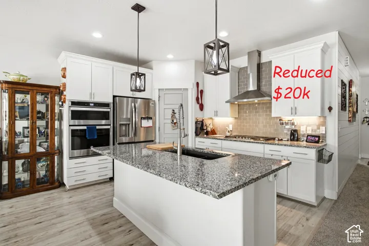 Kitchen featuring white shaker cabinetry, a center island with a stone-finish countertop and integrated sink, stainless steel appliances, a brick-style tile backsplash, and two geometric-frame pendant lights