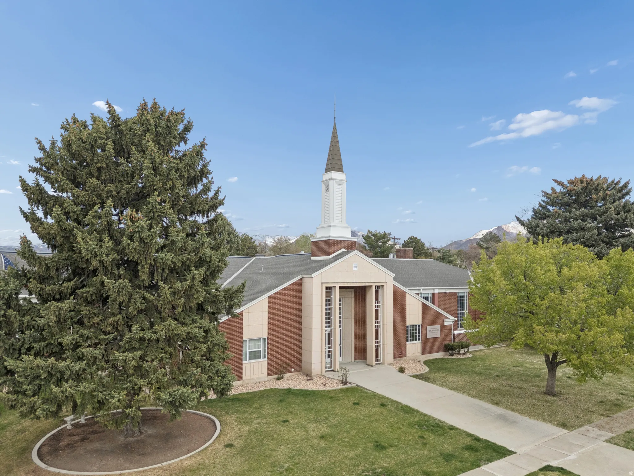 Brick exterior with a prominent steeple, paved walkways, mature trees, and a landscaped front area