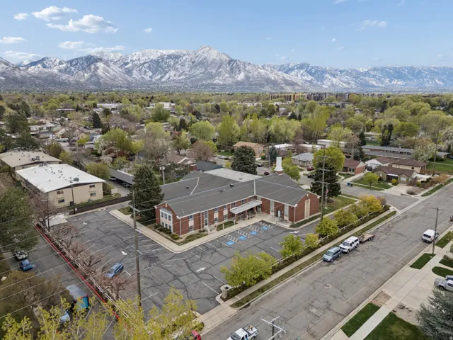 Brick exterior building with gabled roof lines, featuring an expansive parking lot, surrounded by a residential neighborhood with mature trees, and a distant mountain range backdrop