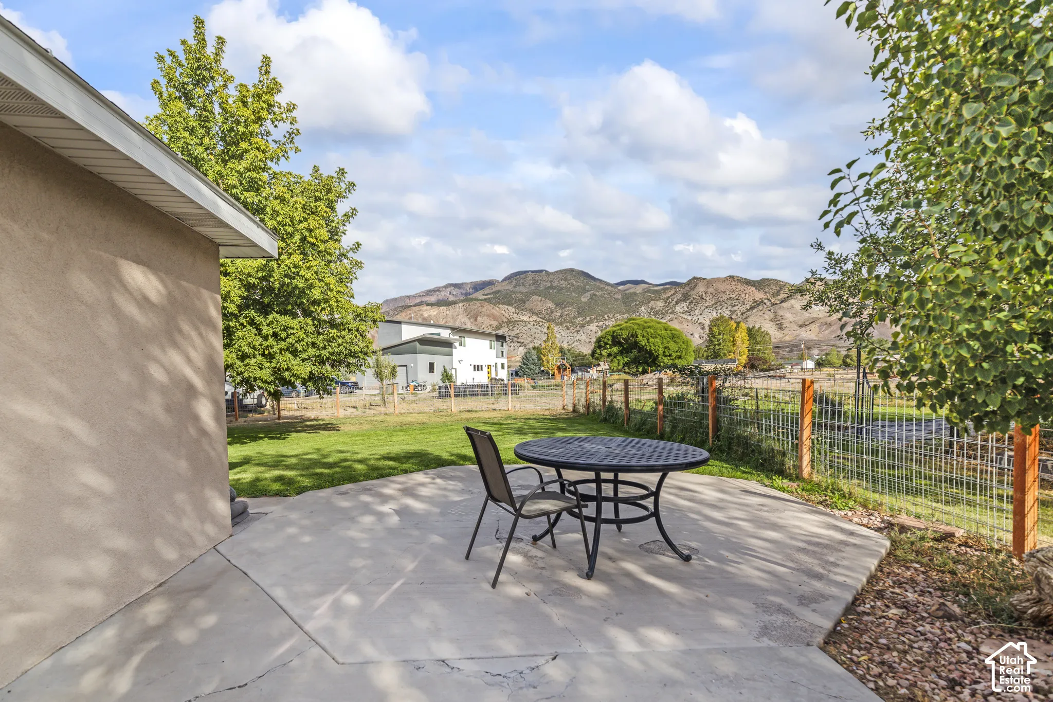 Concrete patio with a wire fence and wood posts, featuring a grass lawn and mature trees, set against a mountain backdrop