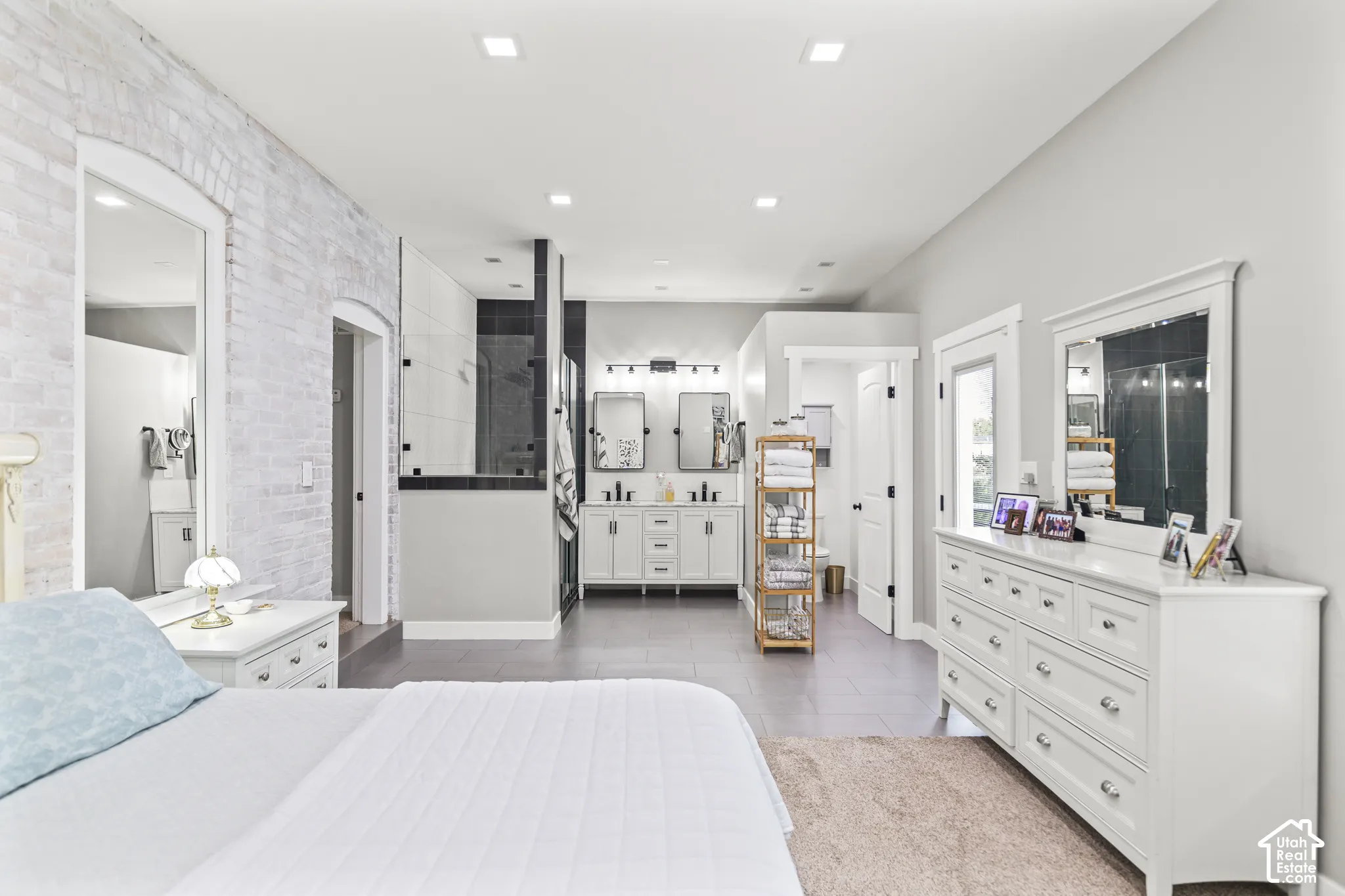 Whitewashed brick accent wall, recessed lighting, dual vanity with undermount sinks, glass-enclosed shower, and tile flooring