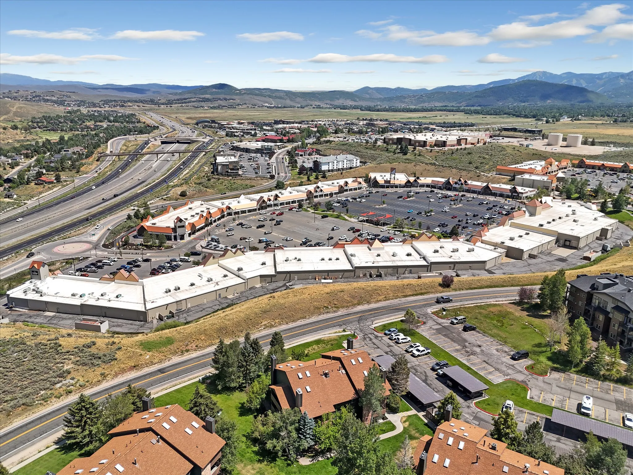 Aerial perspective showcasing a retail complex with extensive parking, adjacent to a multi-lane highway system, and surrounded by mountainous terrain