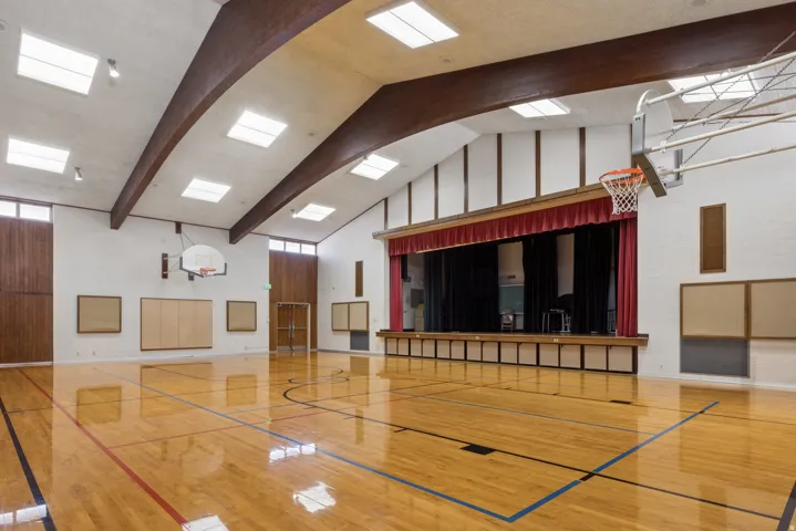 Glossy wood-finish flooring with court markings, an elevated stage with red curtains, white walls, and exposed wood beams