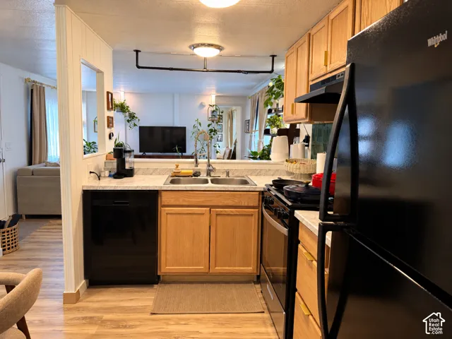 Kitchen featuring light wood cabinetry, black appliances, a double basin sink, light-toned countertops, and wood-finish flooring