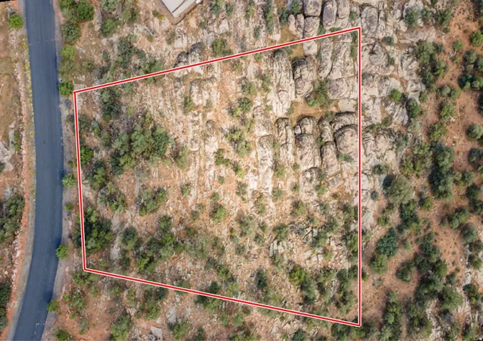 Expansive land parcel featuring natural rock formations, scattered green foliage, and an adjacent paved road