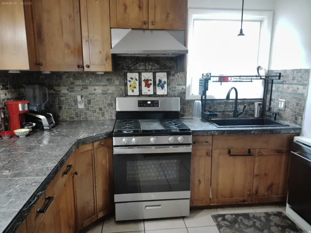 Stainless steel range with a built-in oven, dark stone-finish countertops, natural wood cabinetry, and a mosaic tile backsplash