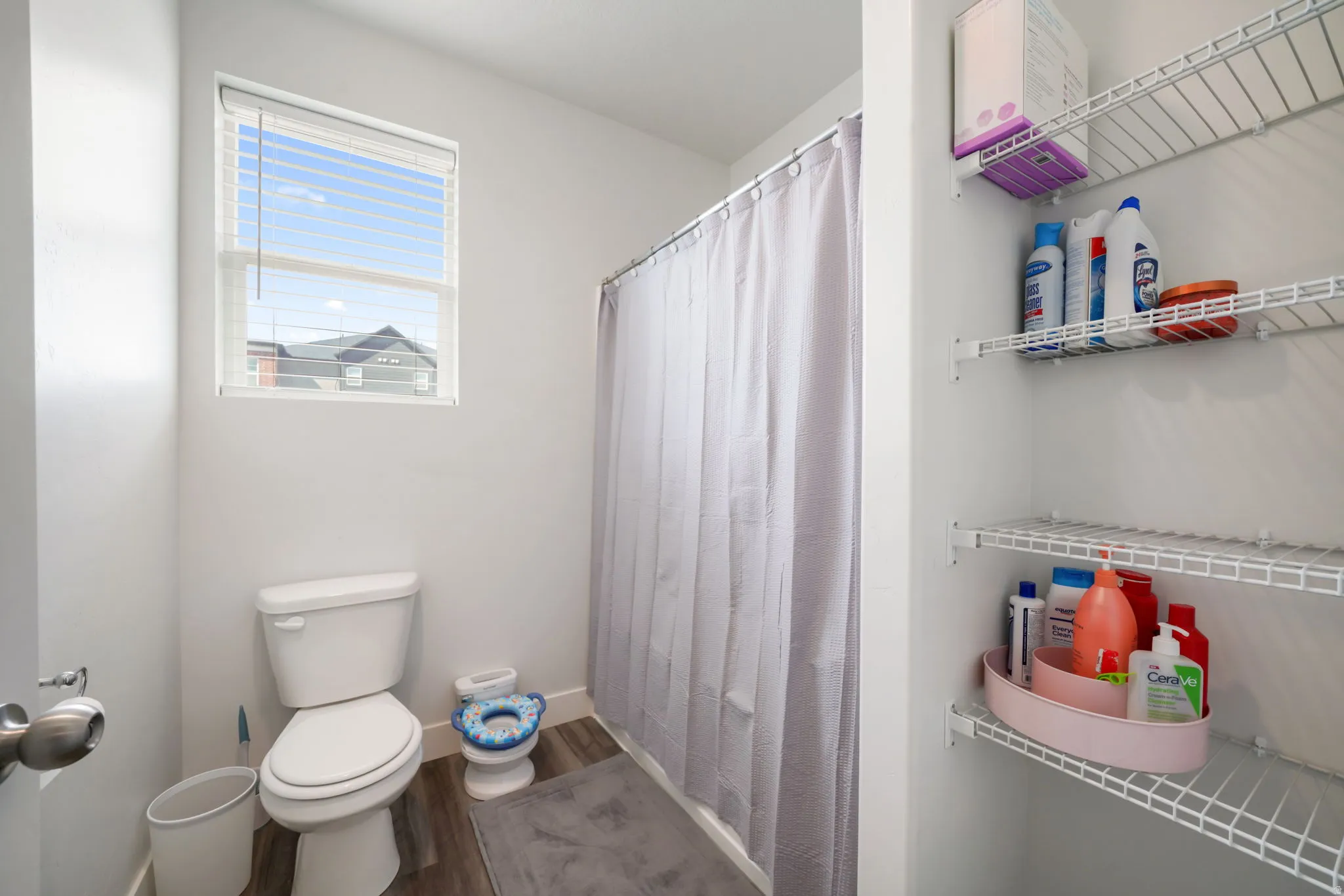 Window with horizontal blinds, wood-finish flooring, white wire shelving, and a built-in shower enclosure