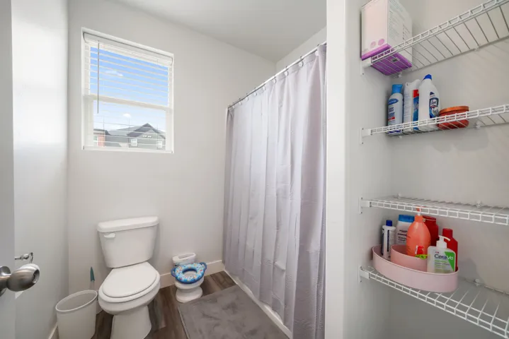 Window with horizontal blinds, wood-finish flooring, white wire shelving, and a built-in shower enclosure