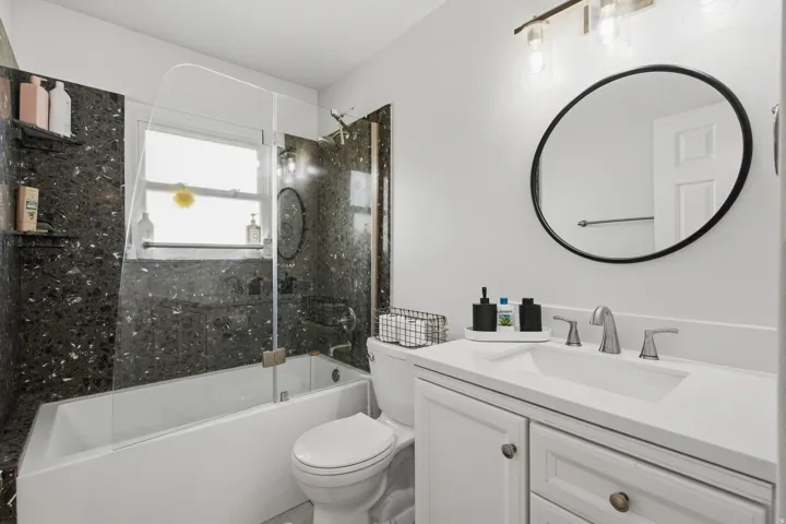White vanity with integrated sink, modern circular mirror, brushed nickel faucet, and a combination tub/shower with dark speckled tile surround