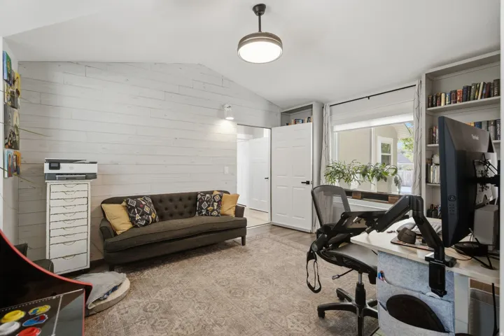 Vaulted ceiling with a flush-mount light fixture, shiplap accent wall, built-in shelving, and a window with roller shades
