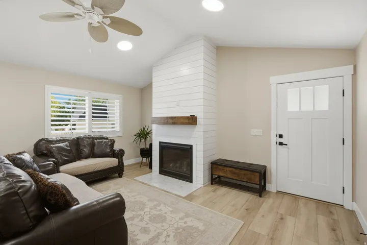 Vaulted ceiling with a ceiling fan, white shiplap fireplace with wood mantel, tile fireplace surround, wood-finish flooring, and a white entry door with glass panels