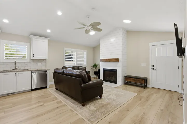 Vaulted ceiling with a ceiling fan, shiplap fireplace with a wood mantle, wood-finish flooring, white cabinetry with a tile backsplash, and stainless steel appliances