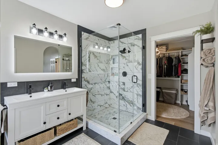 Double vanity with undermount sinks, frameless glass shower enclosure, marble-style shower tile, custom closet shelving, and dark tile flooring