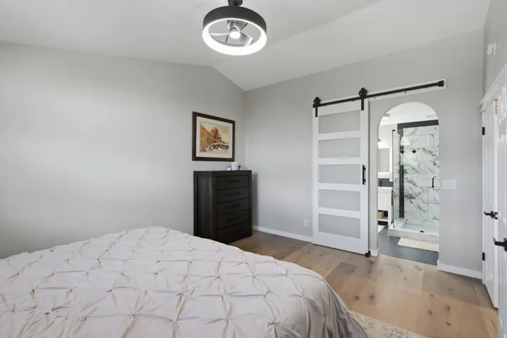 Vaulted ceiling with modern light fixture, wood-finish flooring, sliding barn door with frosted panels, arched entryway, and a walk-in shower with marble-look tile