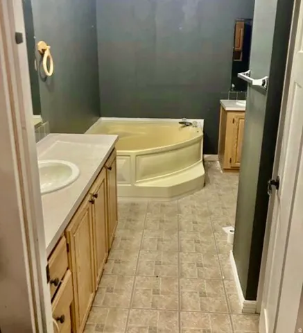 Corner soaking tub, dual vanities with integrated sinks, light wood cabinetry, and tile flooring