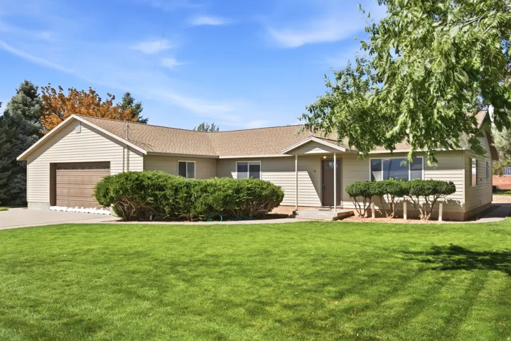 Ranch-style residence featuring light-colored siding, an attached garage with a wood-finish door, a front lawn, and mature landscaping