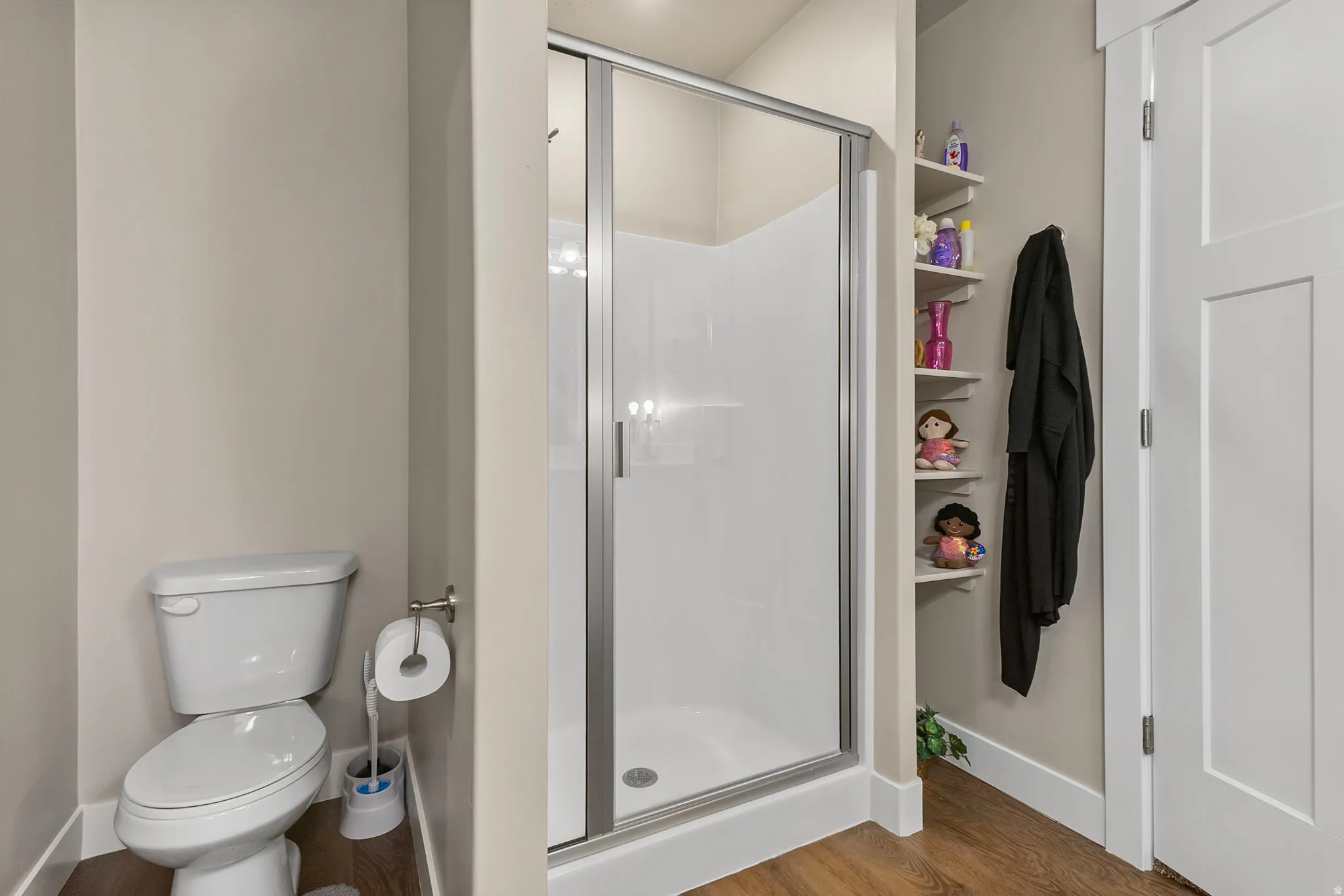 Compact bathroom featuring a white toilet, enclosed shower with sliding glass door, wood-finish flooring, and built-in corner shelving