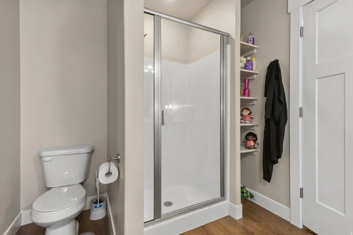 Compact bathroom featuring a white toilet, enclosed shower with sliding glass door, wood-finish flooring, and built-in corner shelving