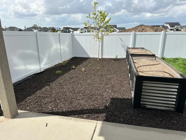 Backyard area featuring a white privacy fence, mulched garden beds, a young tree, and two raised planters with irrigation