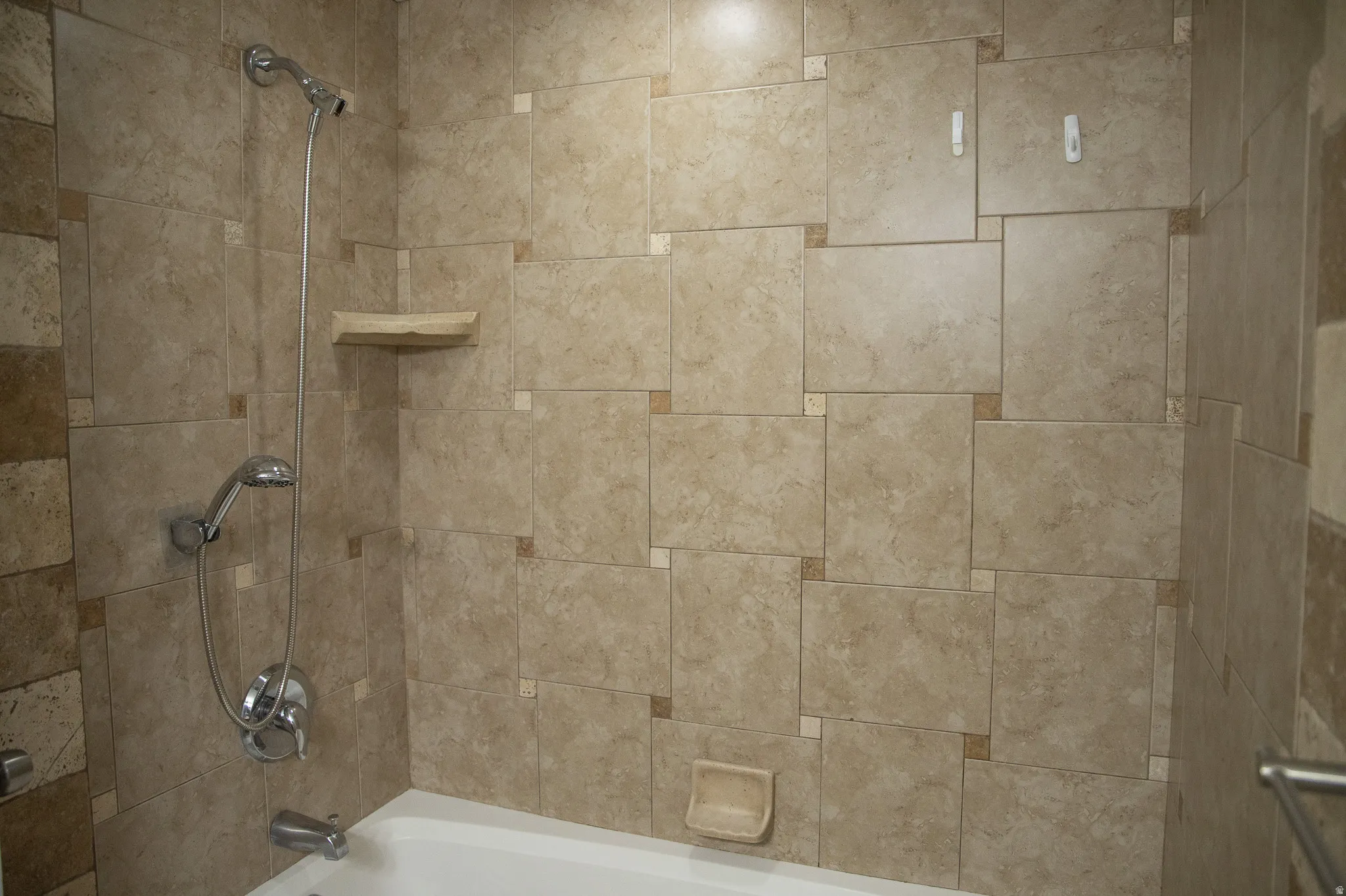 Bathtub with a handheld shower fixture, built-in corner shelf, and textured tile surround