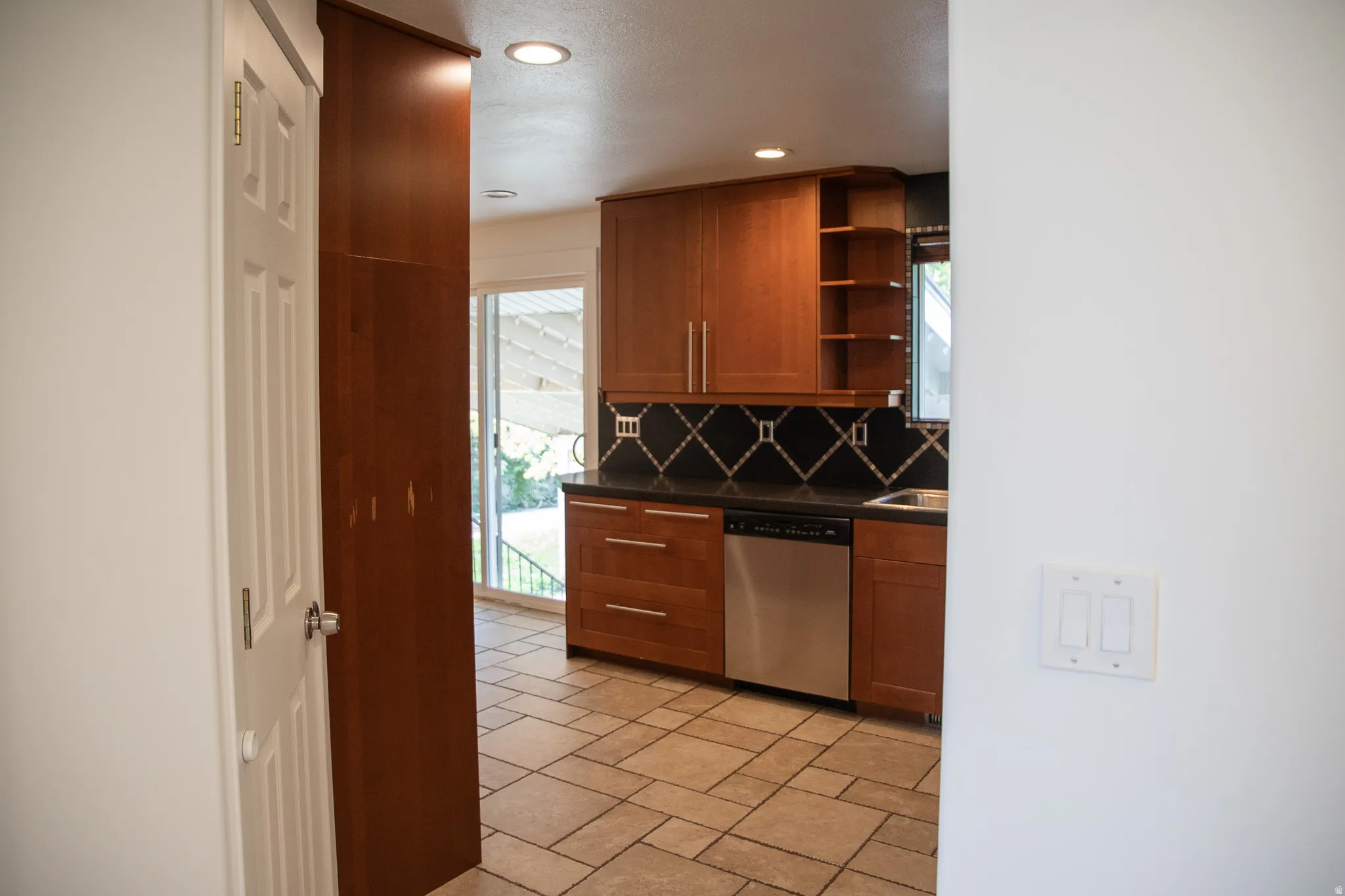 Wood cabinetry, tile flooring, black countertops, patterned backsplash, and stainless steel dishwasher