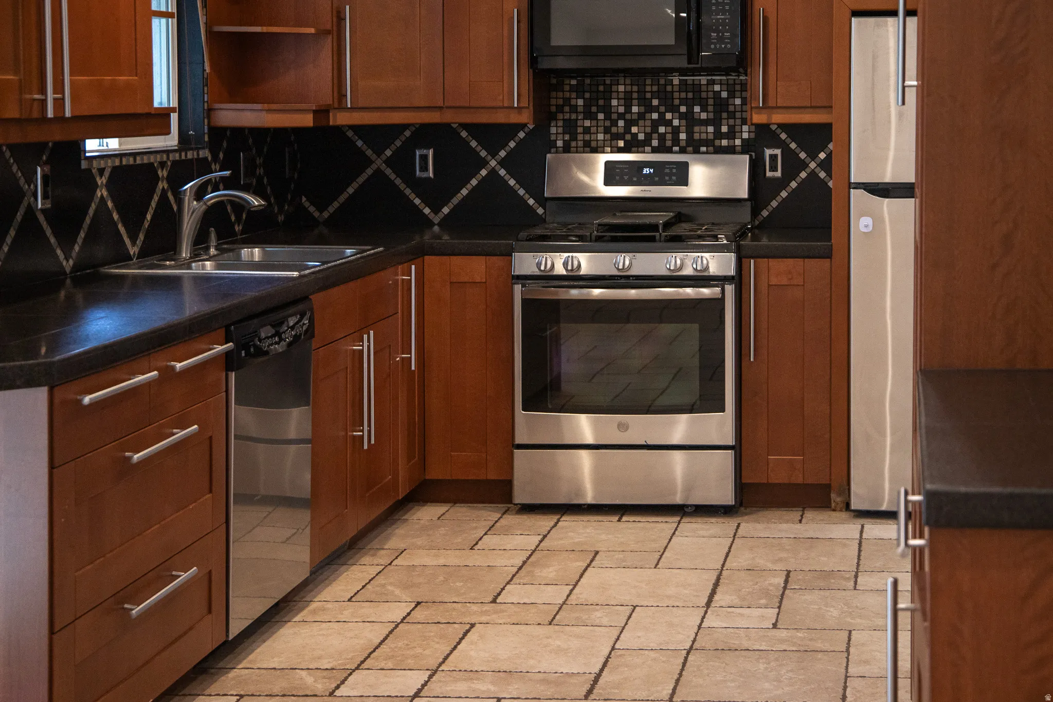 Kitchen featuring wood-finish cabinetry, stainless steel appliances, dark countertops, a dark tile backsplash with diamond accents, and light-toned floor tiling