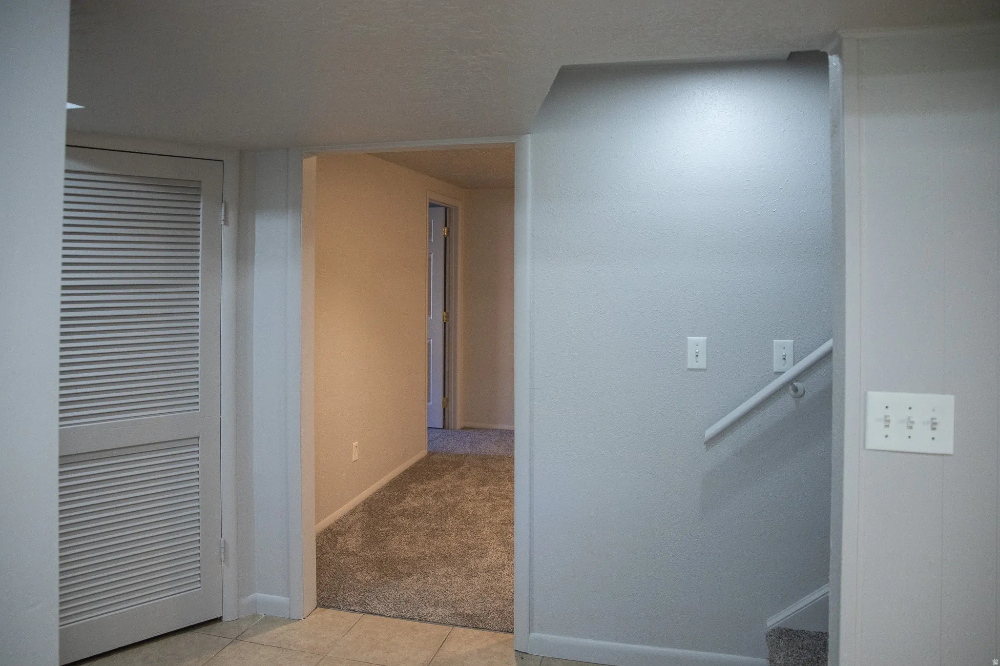 Carpeted flooring extending through a doorway, tiled flooring, louvered door, painted walls, and a staircase with a handrail