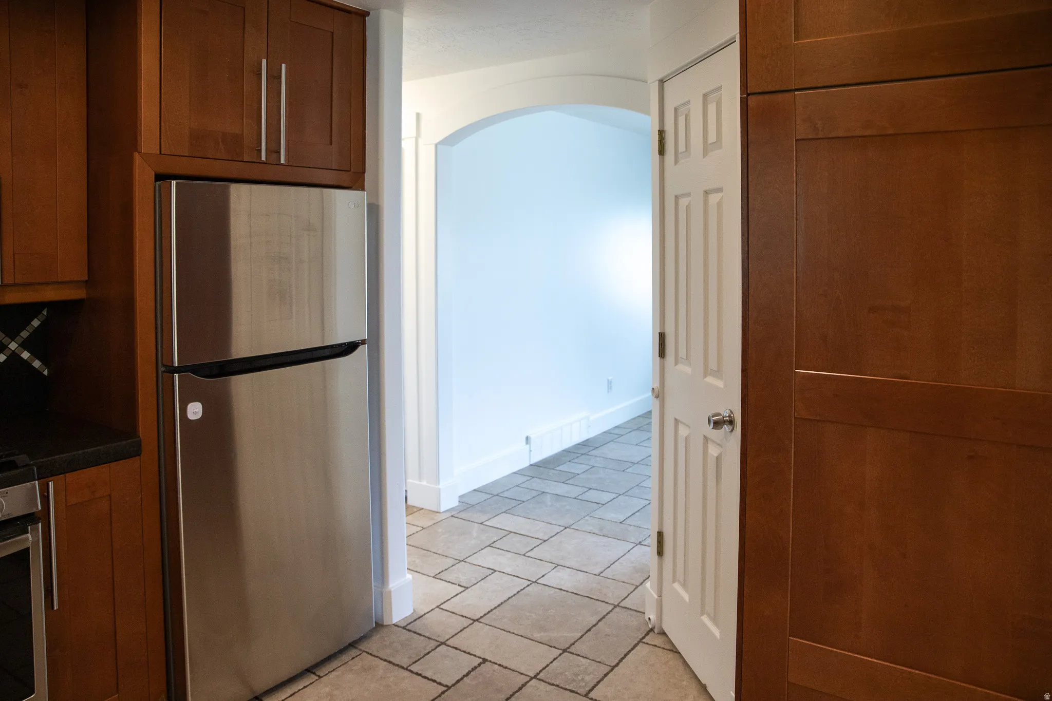Stainless steel refrigerator, wood-finish cabinetry, tile flooring, arched doorway, and a paneled interior door