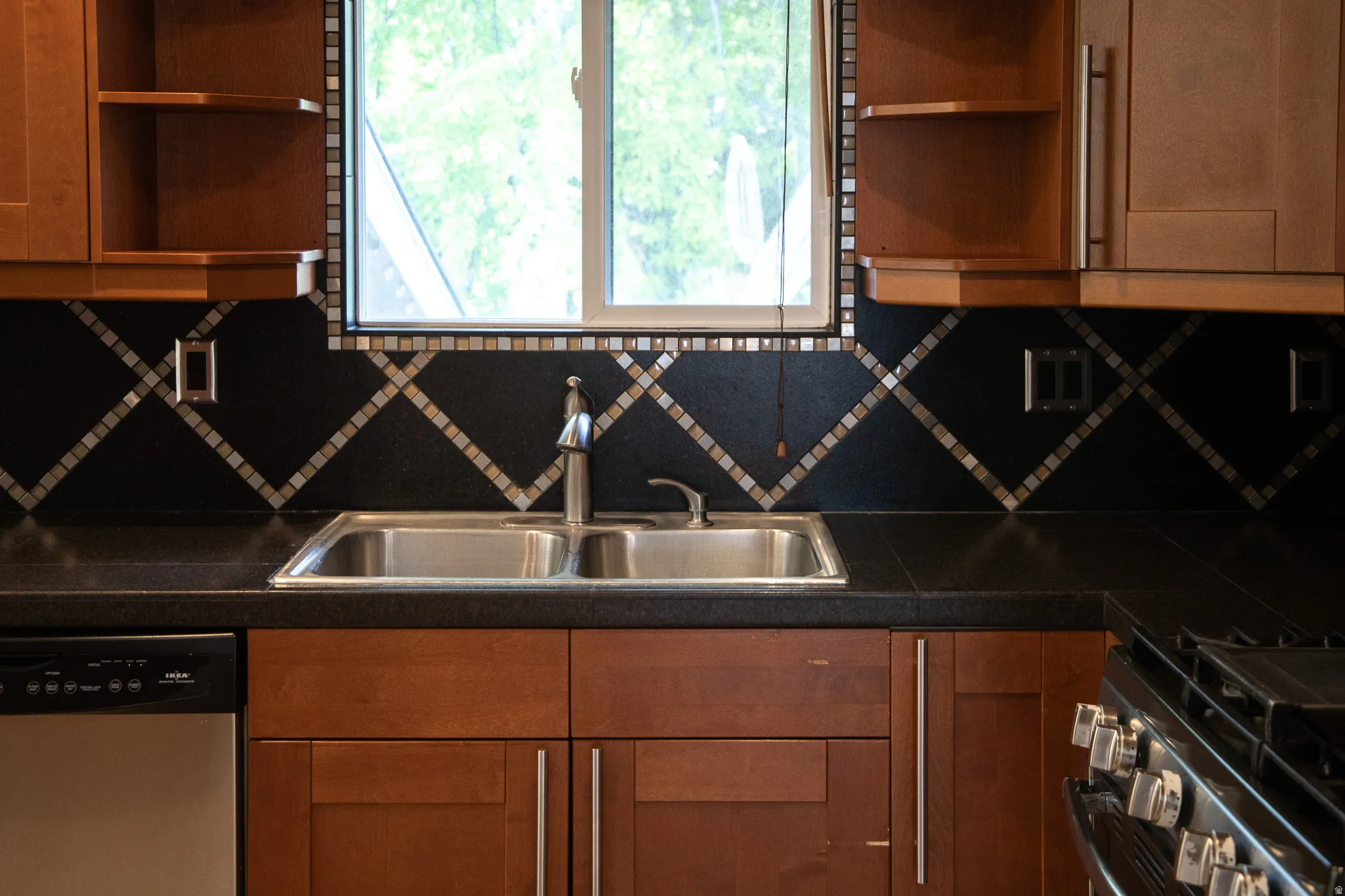 Wood-finish cabinetry, black countertops, stainless steel double basin sink, and a black and metallic tile backsplash with a diamond pattern