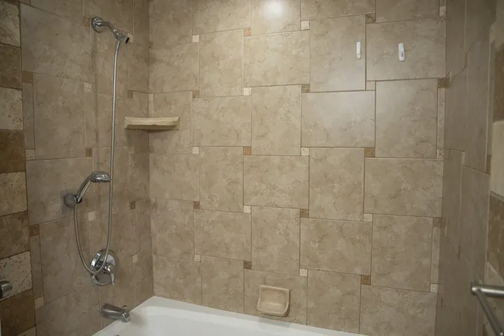 Bathtub with a handheld shower fixture, built-in corner shelf, and textured tile surround