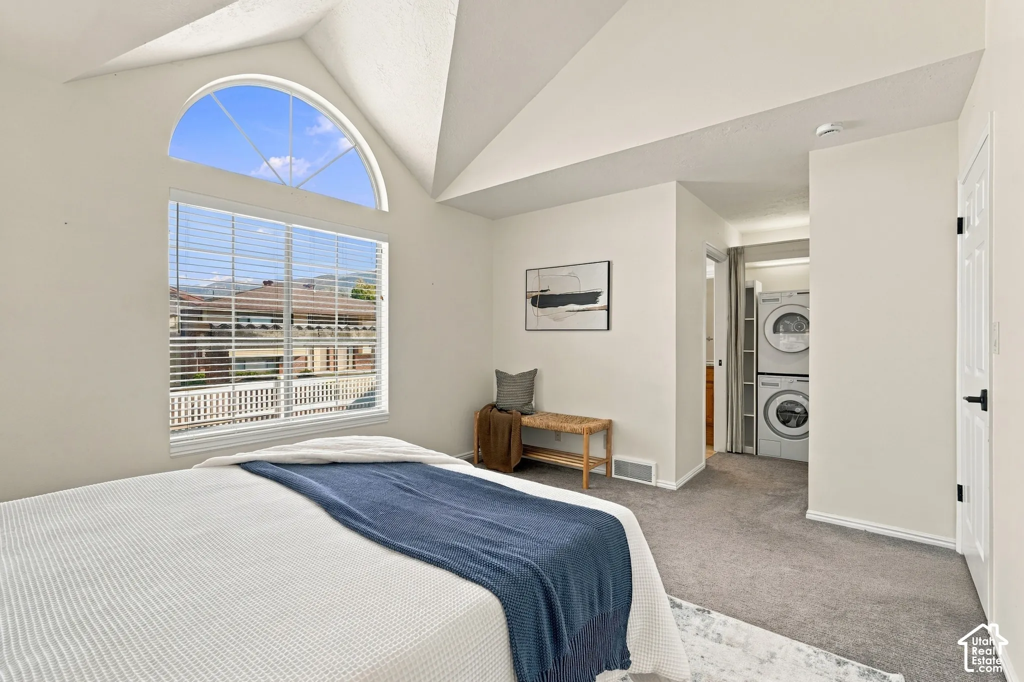 Vaulted ceiling, arched transom window, large window with blinds, carpeted flooring, and stacked laundry appliances