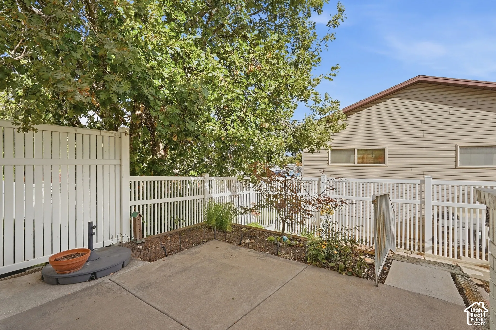 Private concrete patio area featuring white fencing, a raised garden bed, and mature trees