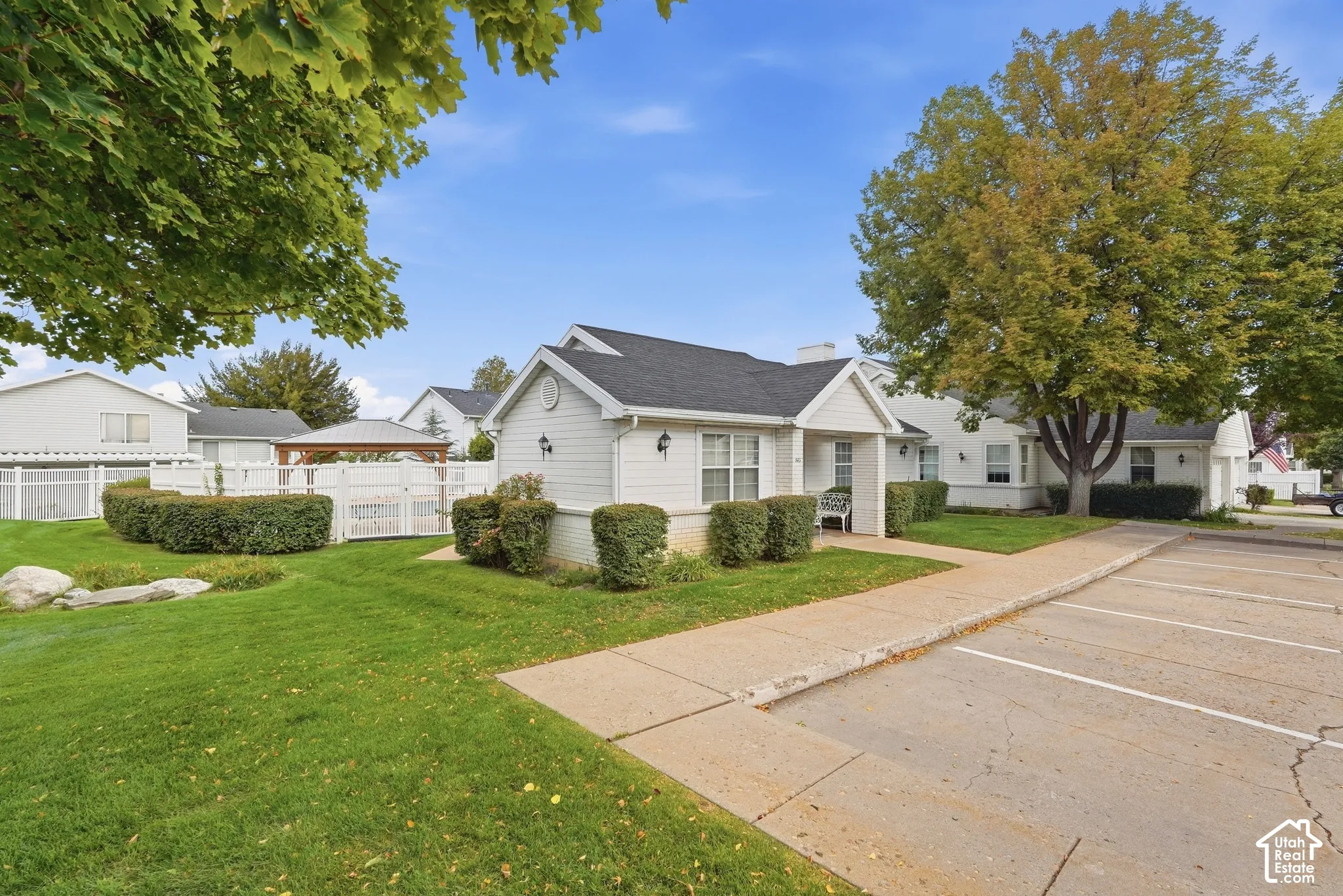 White siding exterior with a dark shingle roof, established landscaping, and a paved parking area
