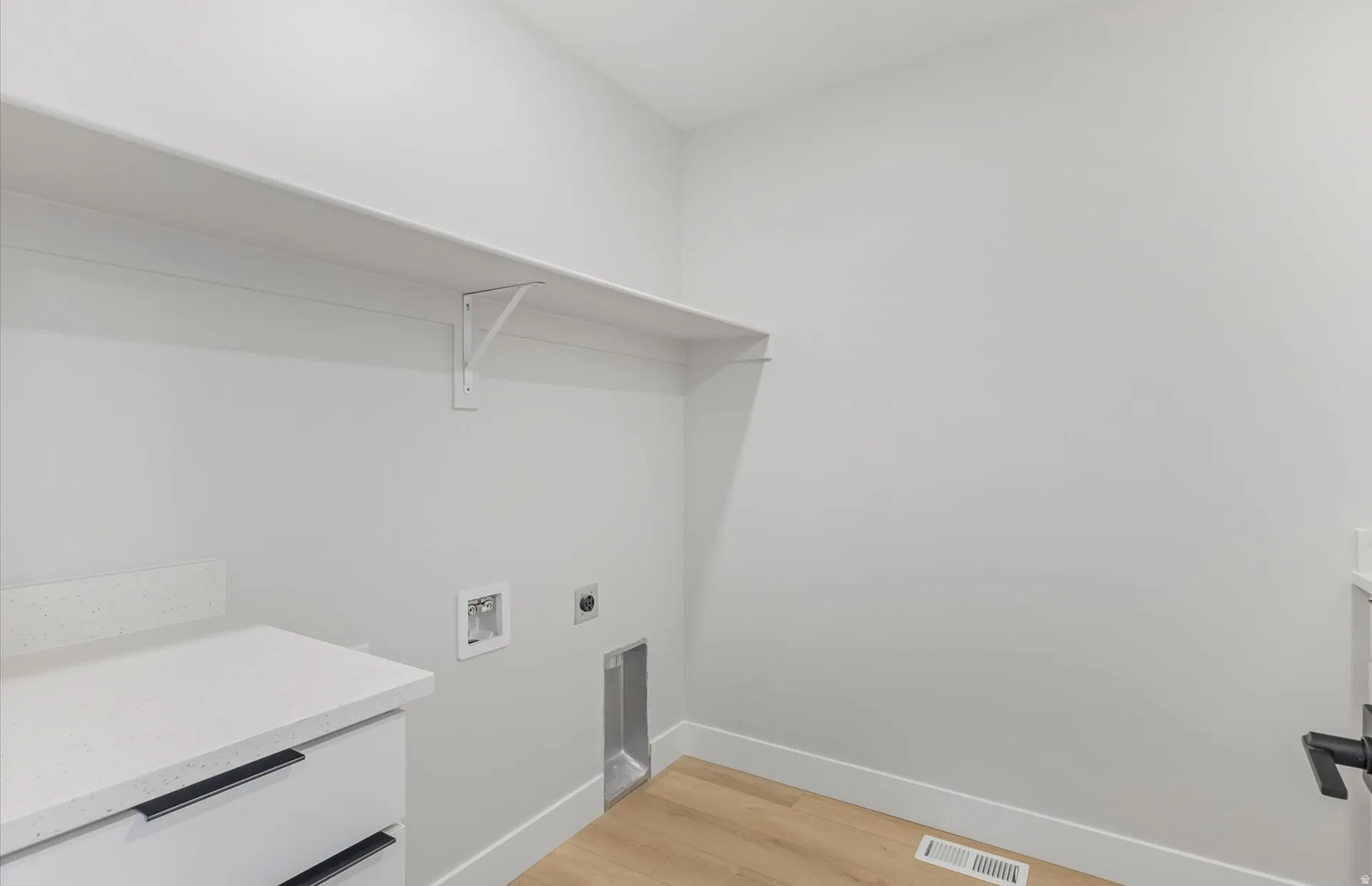 White wall-mounted shelving above appliance hookups, wood-finish flooring, and a built-in cabinet with a light-colored countertop