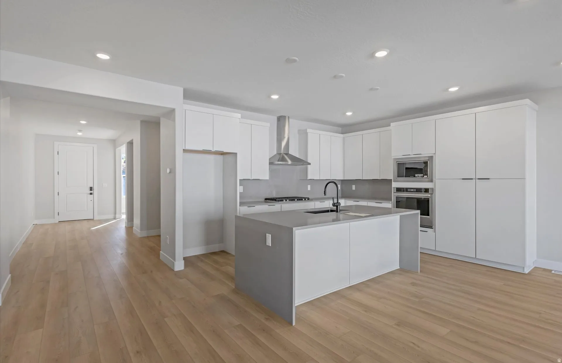 Kitchen featuring white flat-panel cabinetry, a center island with sink, stainless steel appliances, a stainless steel range hood, and wood-finish flooring