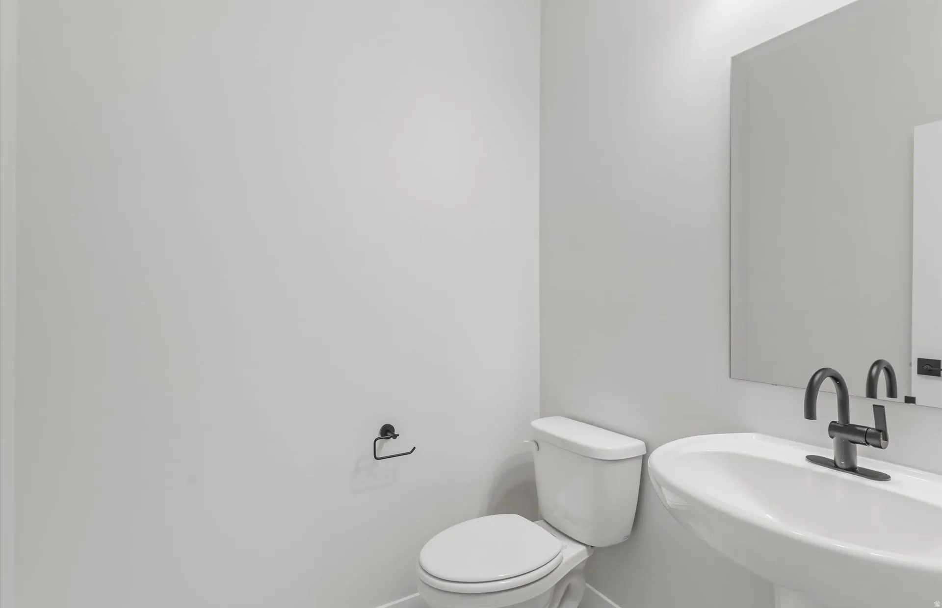 Pedestal sink, matte black faucet, wall-mounted mirror, modern toilet, and white walls