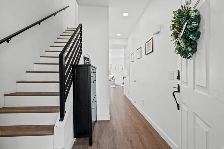 Wood-finish flooring, white walls, recessed lighting, white paneled door with black hardware, and a modern stair railing with wood treads
