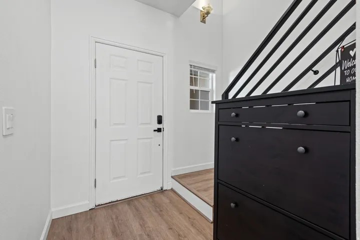 Wood-finish flooring with a white paneled door, small multi-pane window, and an adjacent stairway with black railing