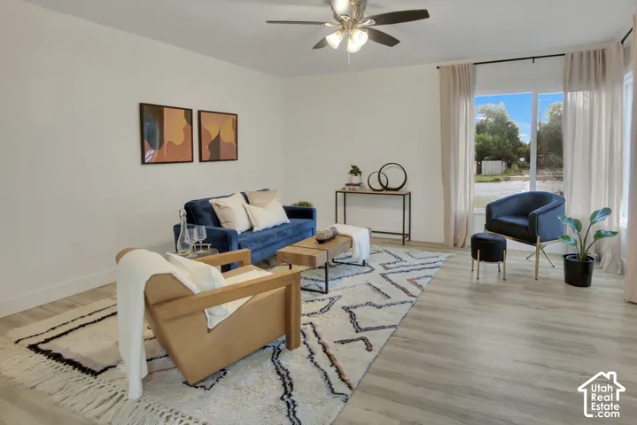 Open-concept living area featuring wood-finish flooring, a ceiling fan with integrated lighting, and large windows with privacy curtains
