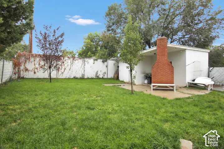 Expansive backyard featuring a manicured lawn, a red brick outdoor fireplace, a white privacy fence, and mature trees