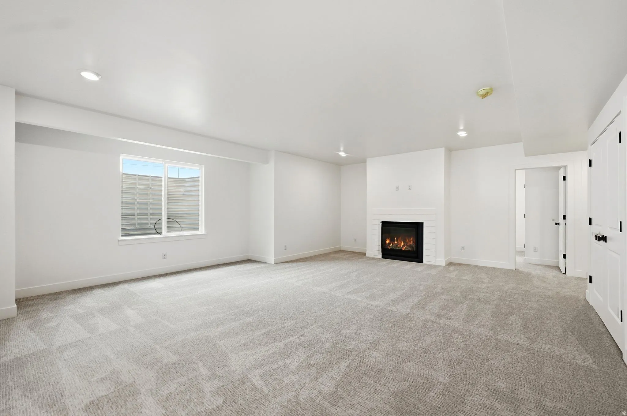 Expansive room featuring light-toned carpeting, a gas fireplace with a white tile surround, recessed lighting, and a window providing natural illumination