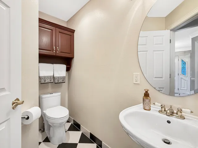 Pedestal sink with polished chrome faucet, round frameless mirror, dark wood-finish wall-mounted cabinetry, and black and white checkered floor tiling